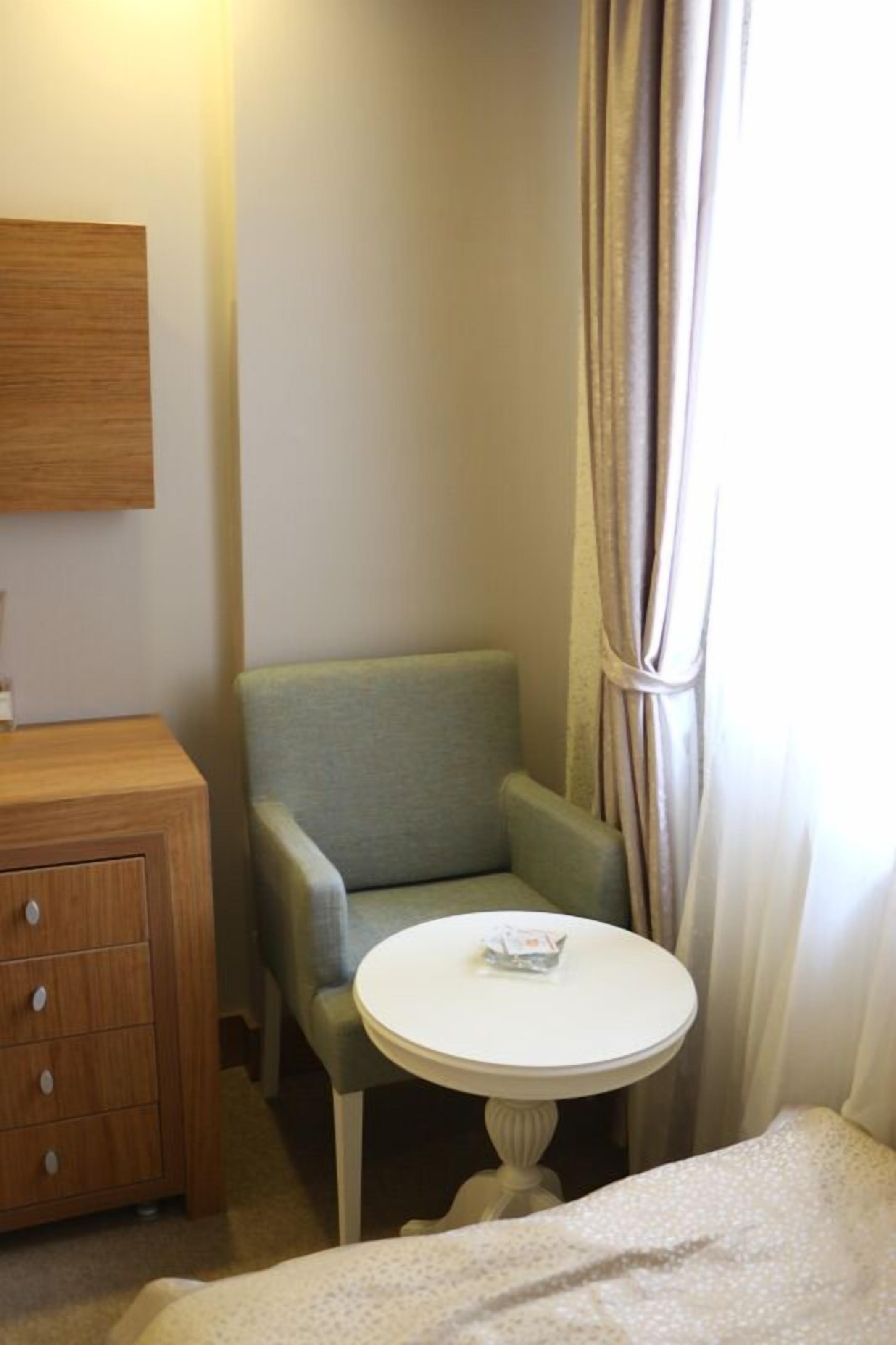 Una habitación pequeña con un aparador de madera, un sillón verde, una mesa redonda blanca y una ventana con cortinas.