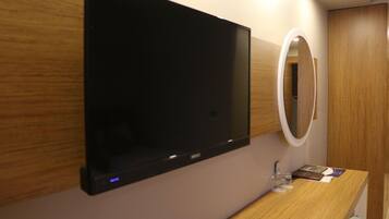 Quarto de hotel moderno com televisão de tela plana na parede, penteadeira de madeira com espelho e uma pequena mesa com um copo de água e um livro.