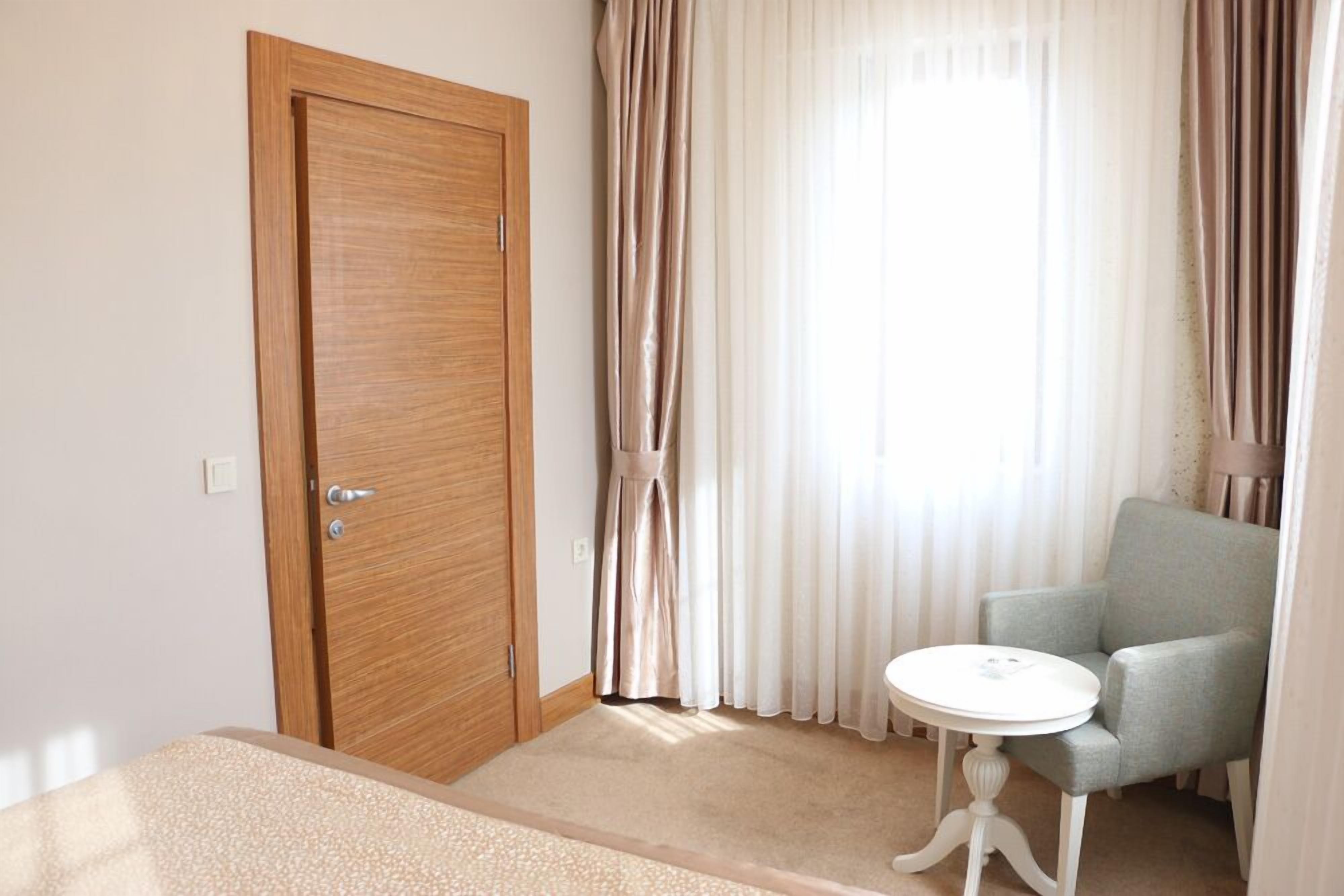 Una habitación con puerta de madera, una mesita redonda pequeña, un sillón azul claro y una ventana con cortinas traslúcidas.