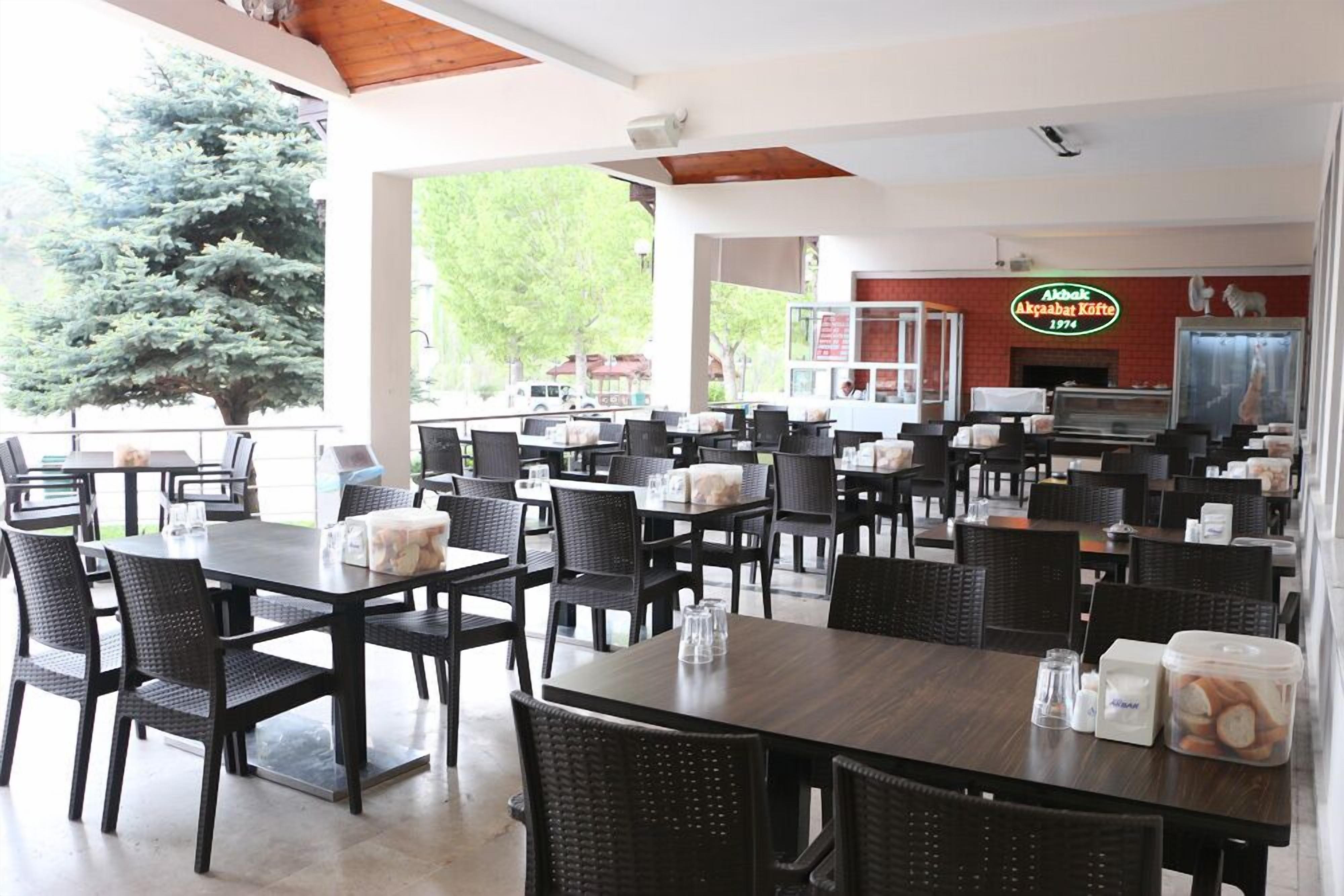 A spacious restaurant with multiple tables and chairs, a large tree visible through the windows, and a sign indicating the establishment's name and year.