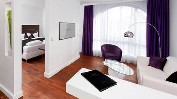 A modern hotel room with a white sofa, a glass coffee table, a purple armchair, and a bed with white bedding.