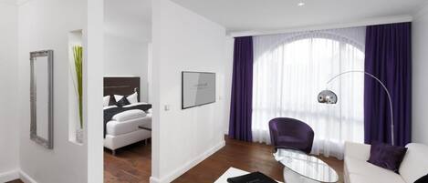 A modern hotel room with a white sofa, a glass coffee table, a purple armchair, and a large window with purple curtains.