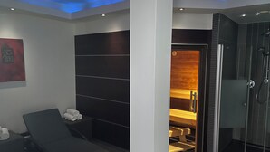 Une salle de spa moderne équipée d’une table de massage, d’un sauna et d’un espace de douche clos par des parois de verre.