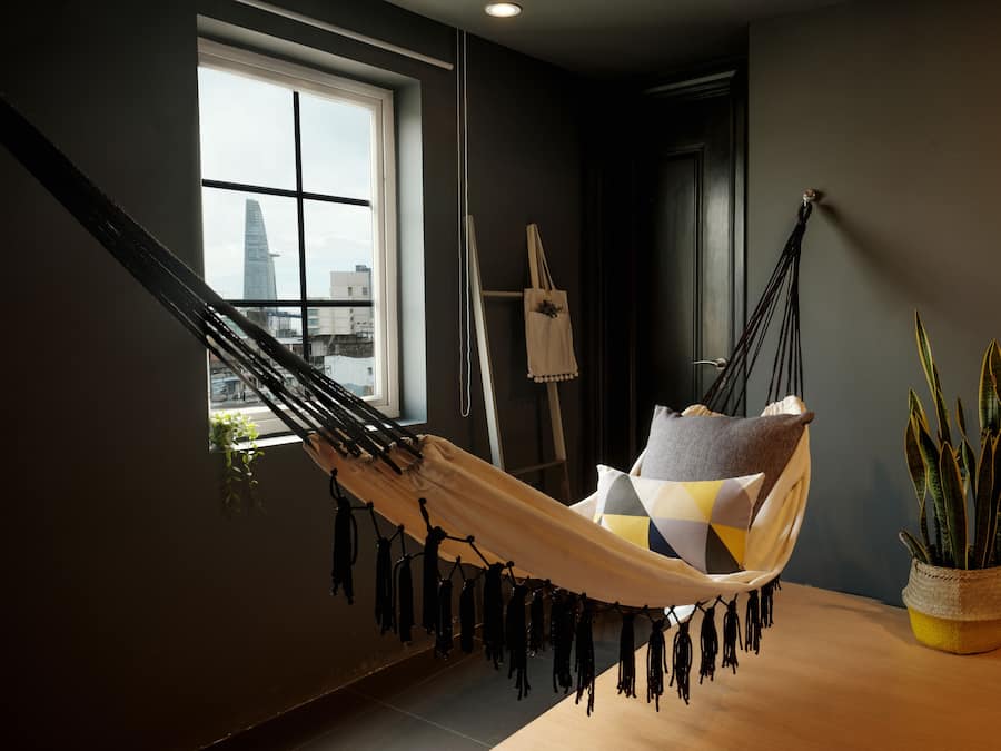 A hammock chair with a view of a cityscape through a window, a potted plant, and a painting on the wall.