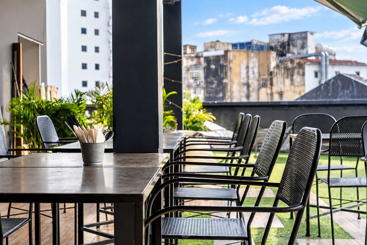 An outdoor patio with a long table and multiple chairs, surrounded by greenery and overlooking a cityscape.