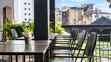 An outdoor patio with a long table and multiple chairs, surrounded by greenery and overlooking a cityscape.