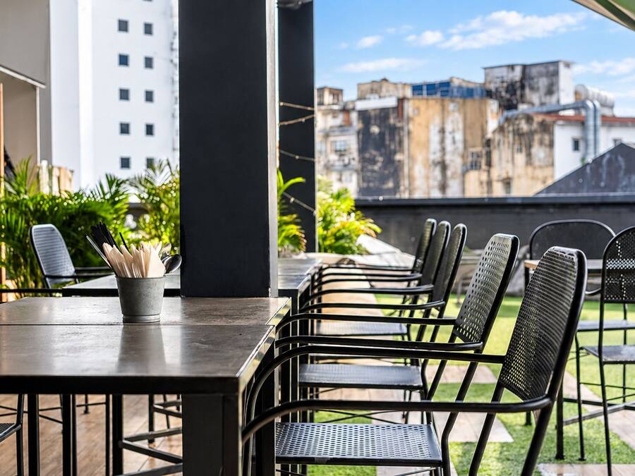An outdoor patio with a long table and multiple chairs, surrounded by greenery and overlooking a cityscape.