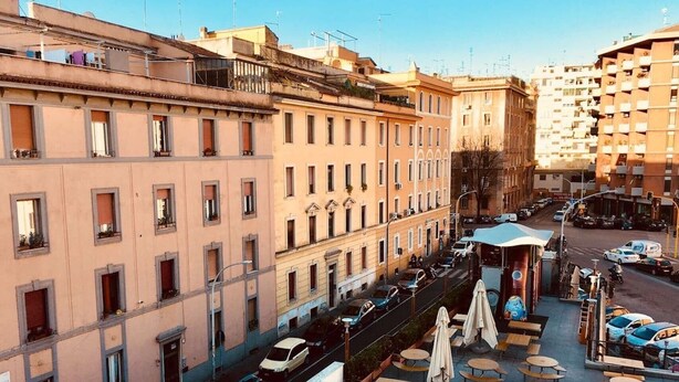 A multi-story building with a row of balconies, a street with parked cars, and outdoor seating area with tables and umbrellas. - Panoramic Castle Apartment, Trastevere, Old Town, Free Parking (Roma)