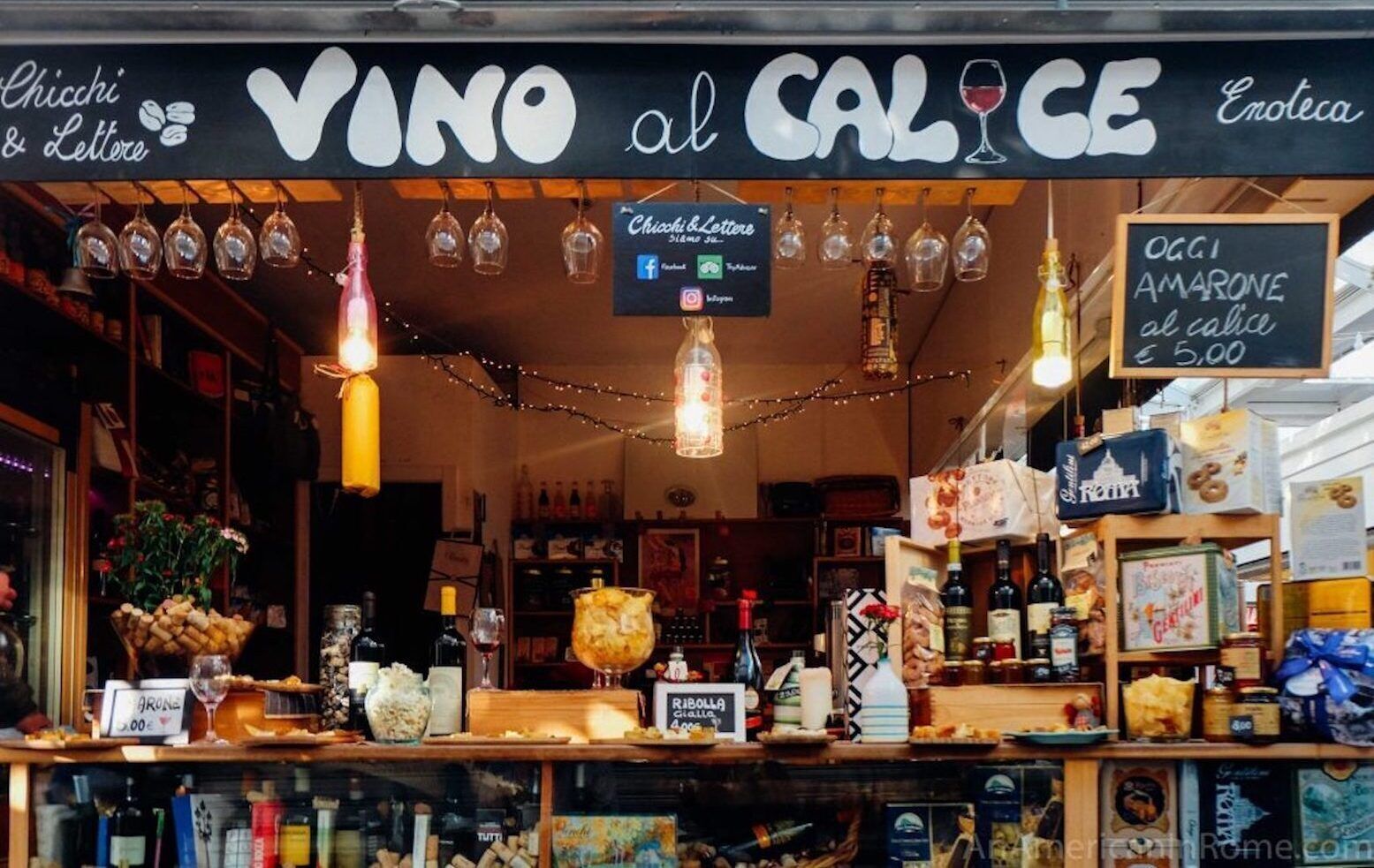 A wine and cheese shop with a sign reading "VINO al Calice Enoteca". There are hanging wine glasses, a variety of bottles and jars on shelves, and a chalkboard sign advertising "OGGI AMARONE al calice €5,00".