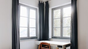 Habitación de esquina con escritorio, silla, ventanas amplias con cortinas y una cama.