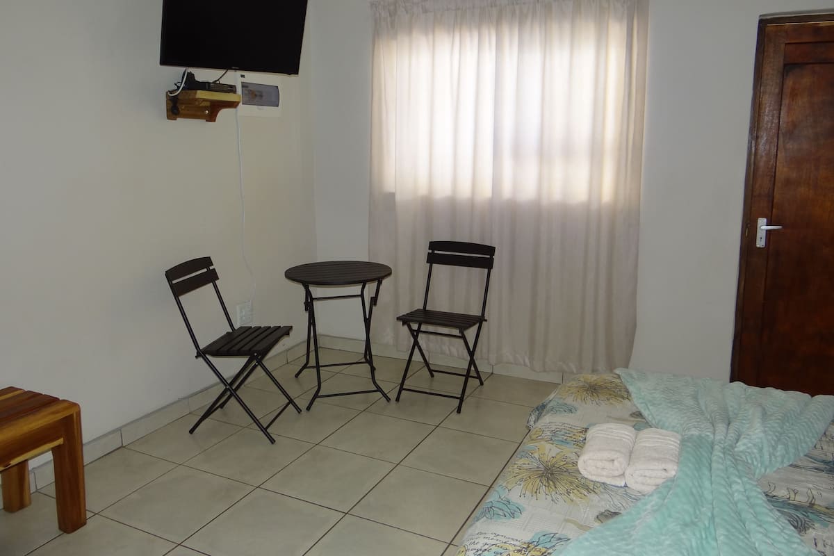 A small, clean room with a bed, two chairs, a table, a TV, and a window with curtains.