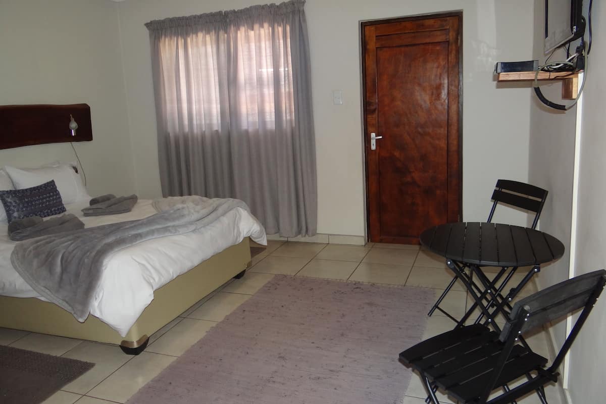 A bedroom with a bed, a wooden door, a small table with chairs, and a wall-mounted TV.