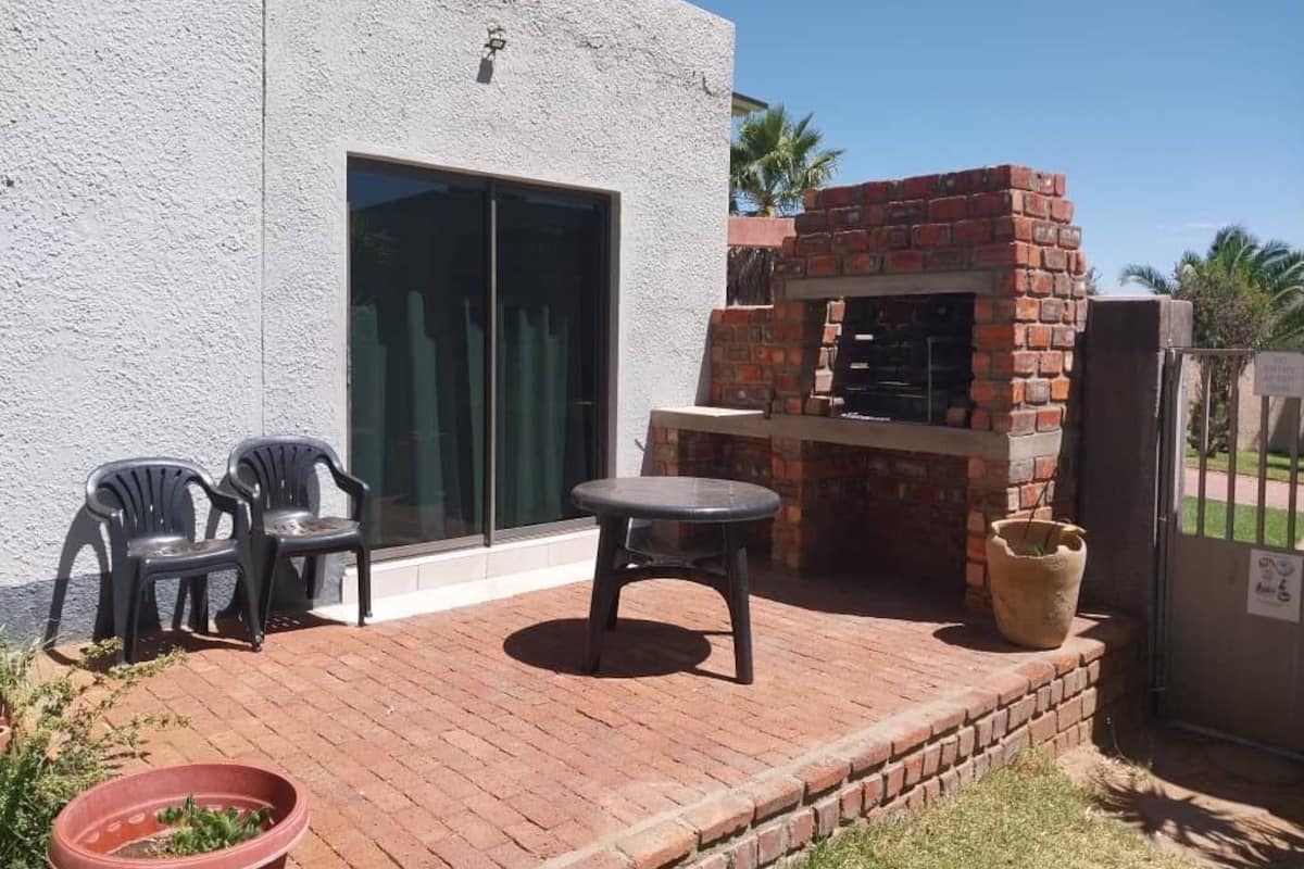 A patio with a brick barbecue, two plastic chairs, a potted plant, and a sliding glass door.