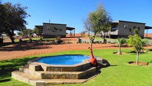 A pool with stone steps, surrounded by grass and trees, with buildings in the background. - Eden Self Catering Chalets (Windhoek)