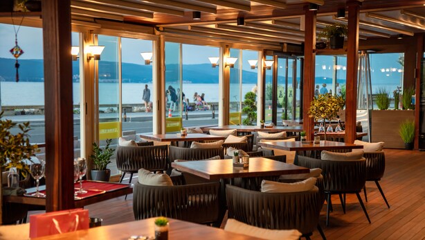 A restaurant with wooden tables and chairs, large windows with a view of the water and mountains, and potted plants. - Paradiso Hotel (Nessebar)