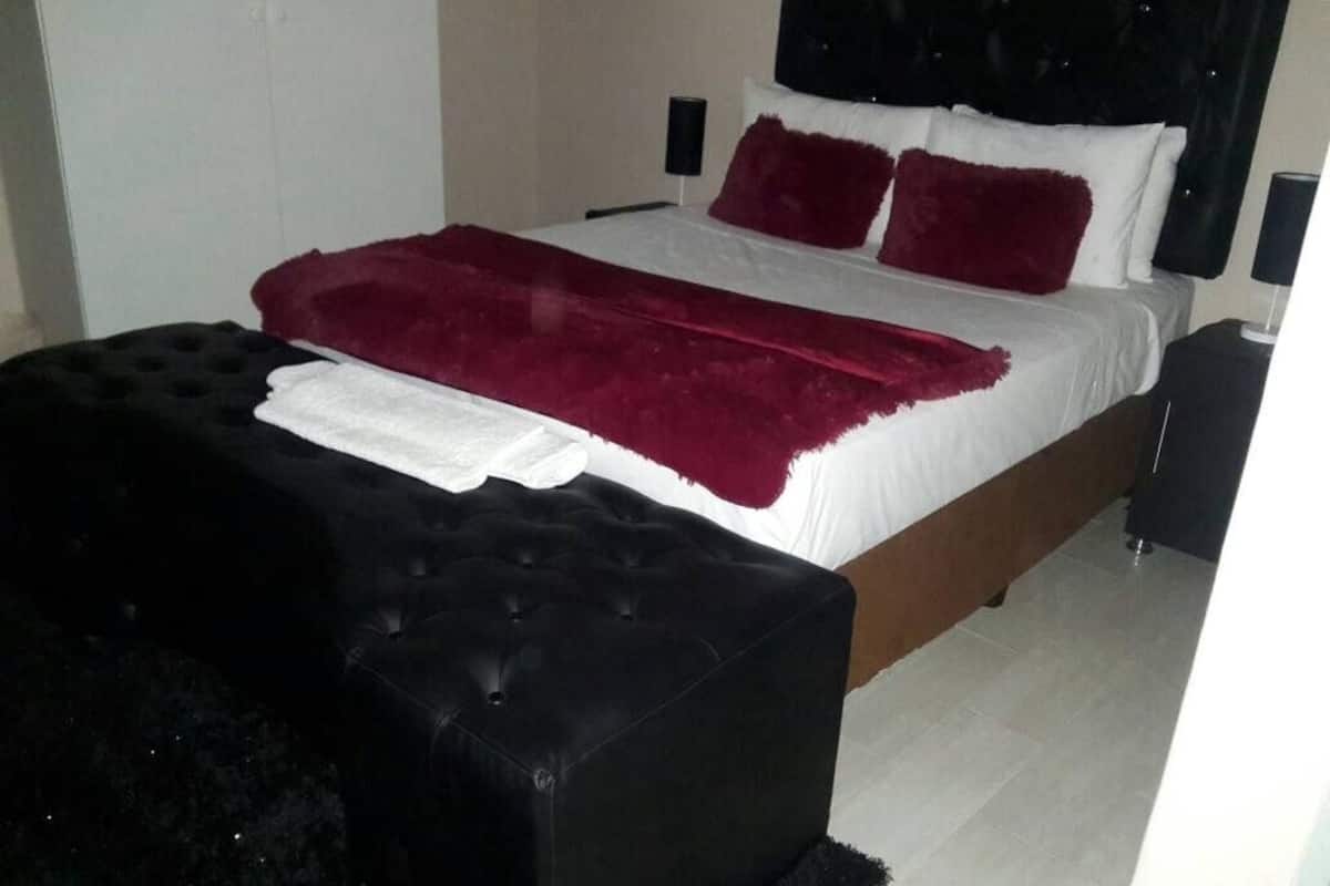A bed with a white and red bedspread, two red pillows, a black ottoman, and a black nightstand with a lamp.