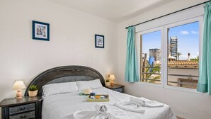 Una cama bien hecha con ropa blanca, una bandeja con alimentos para el desayuno y toallas sobre la cama. Hay dos mesitas de noche con lámparas y una planta pequeña. La habitación tiene un ventanal con vistas a edificios y un balcón.