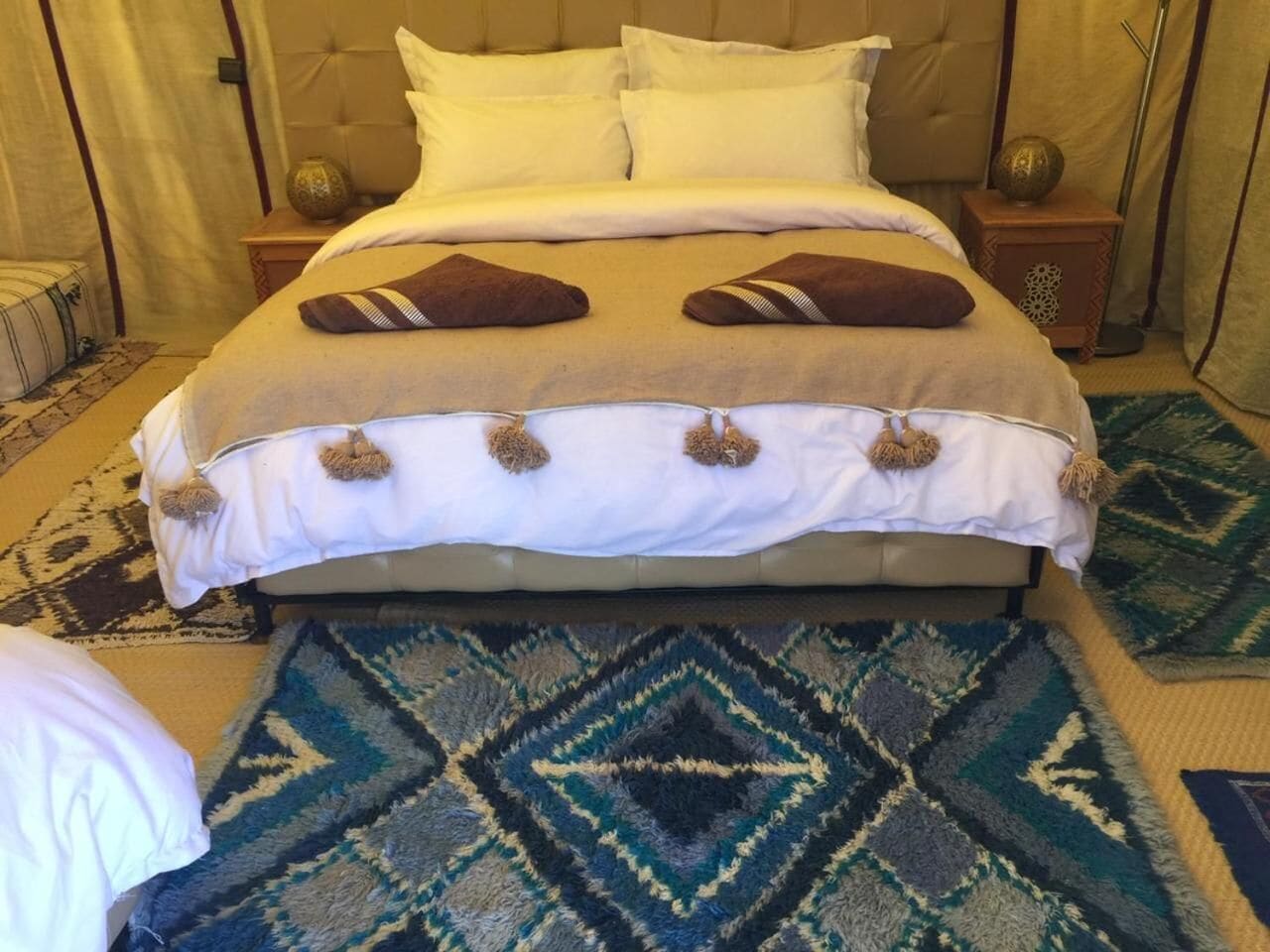 A bed with a beige headboard, white bedding, and two brown pillows. There are two nightstands with decorative items on them. The floor has a patterned rug.