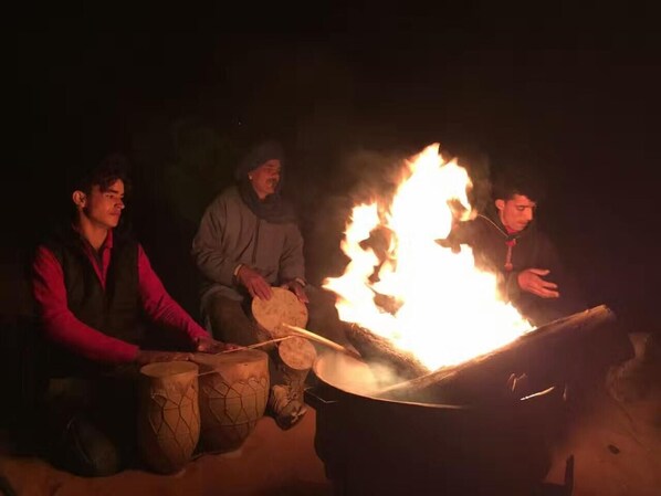 Tres personas alrededor de una fogata, una tocando la batería y otra sosteniendo un objeto plano sobre el fuego.
