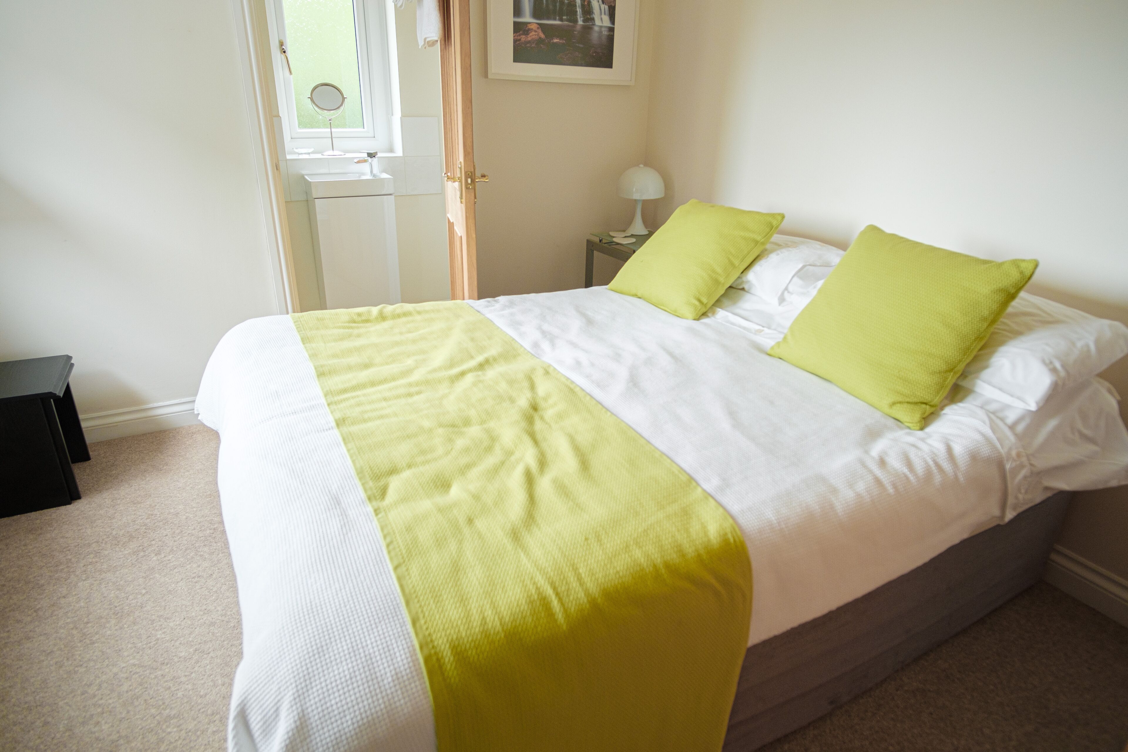 A neatly made bed with a white and yellow bedspread, two green pillows, and a bedside table with a lamp.