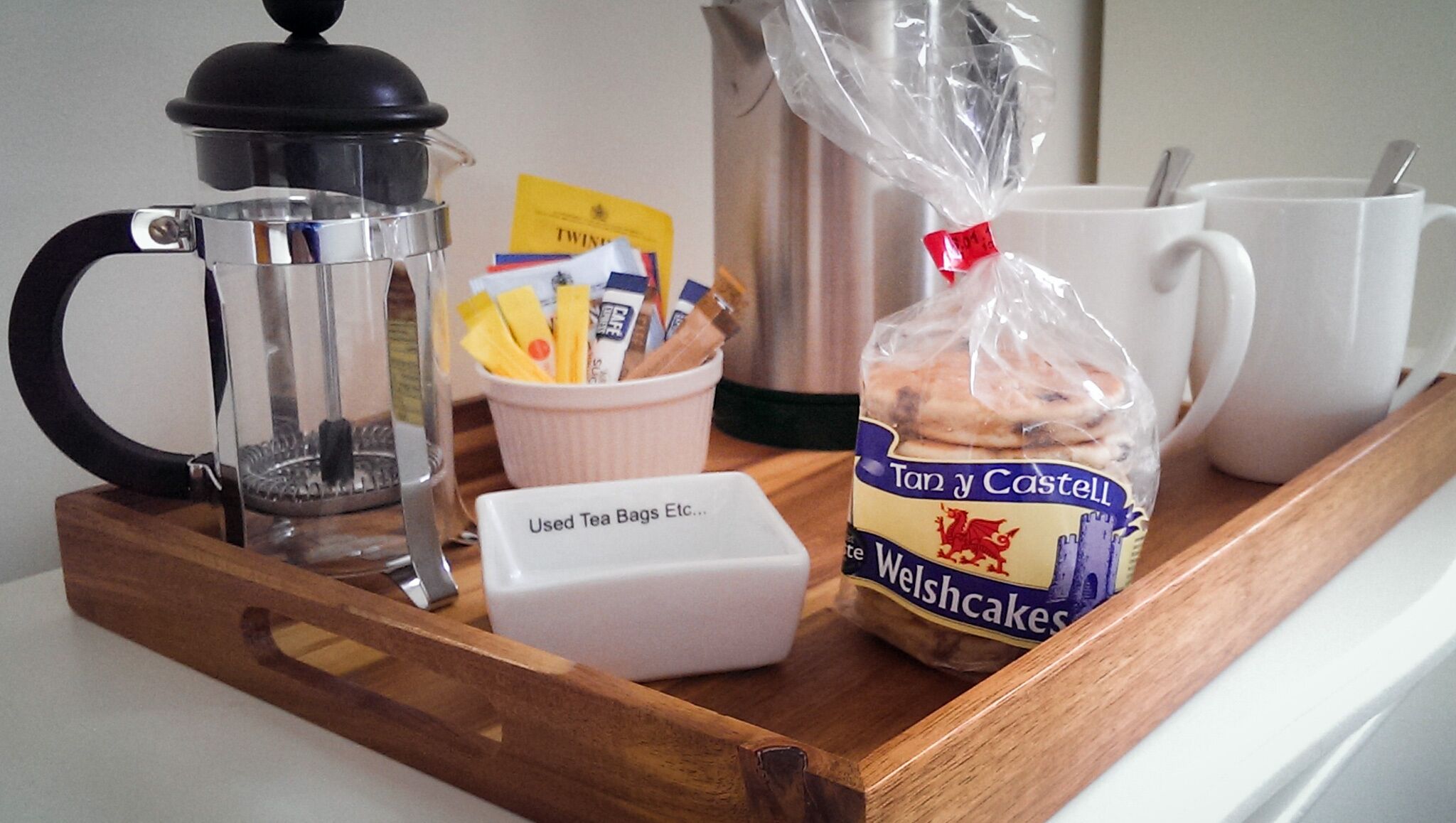 Ett bricka med en kaffebryggare, tepåsar, ett paket walesiska kakor och vita muggar.