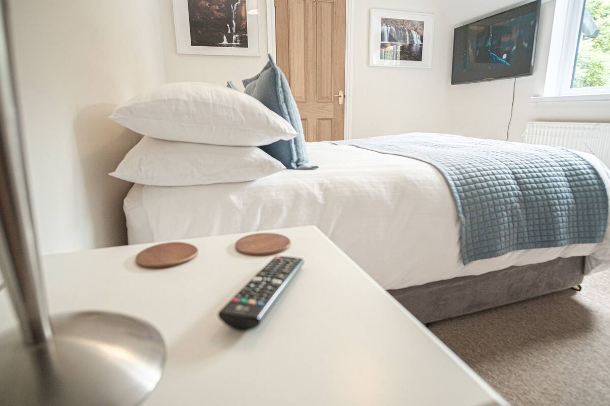 A bedroom with a bed, pillows, a TV, and a remote control on a table.