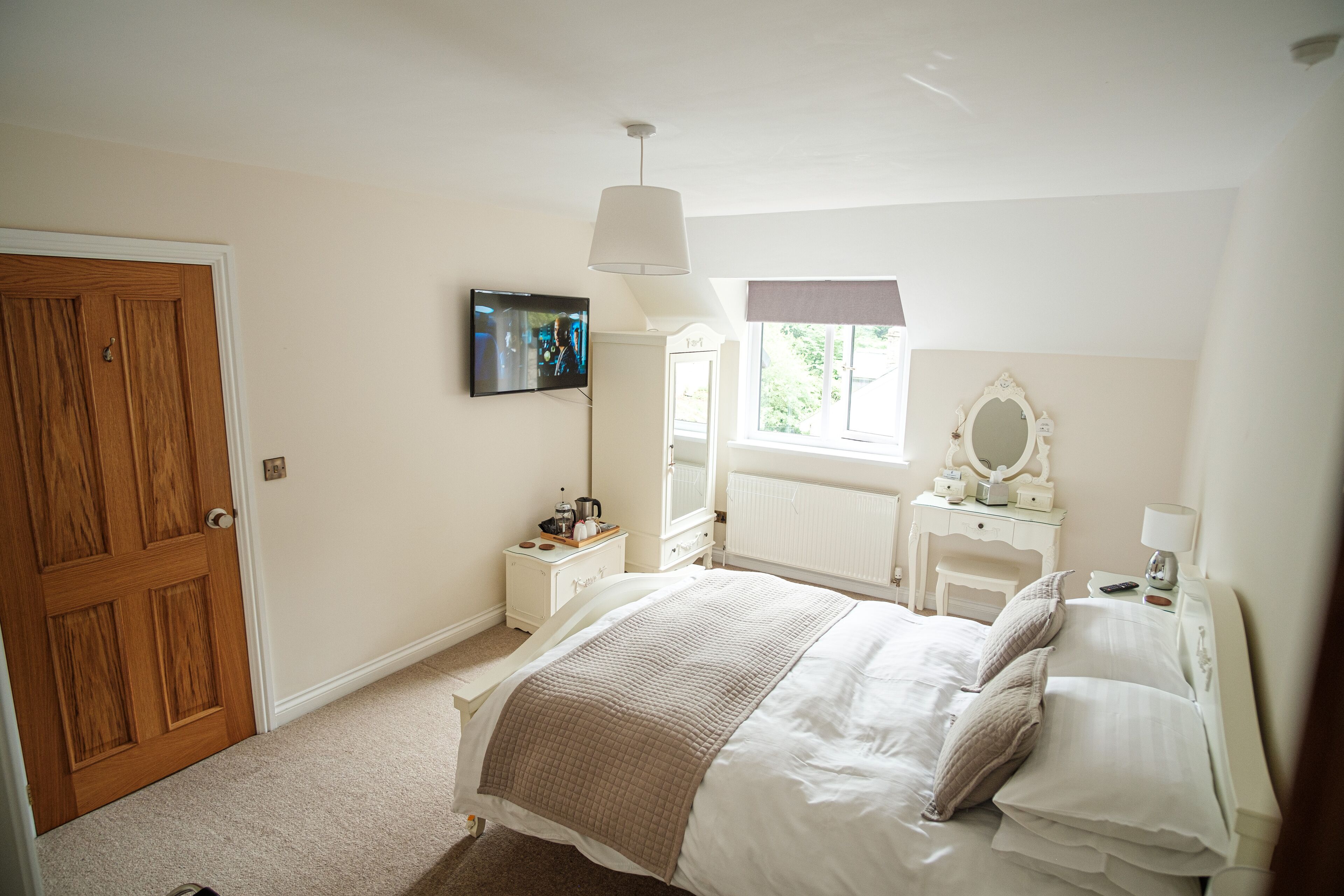Un dormitorio con cama, cómoda, tocador con espejo, televisor en la pared y ventana con vista a follaje.