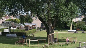 A park with wooden play structures, a tree, and a building in the background. - Vacation home for 10 pers in Westende: Rest on the Coast (Middelkerke)