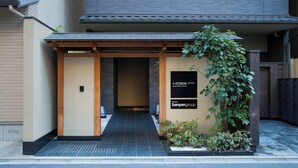 Une entrée de style japonais traditionnel, avec une porte en bois, un petit jardin agrémenté de plantes et une enseigne indiquant le nom de l’établissement.