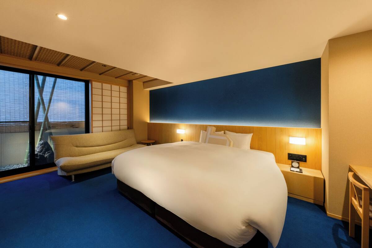 Une chambre d’hôtel avec un grand lit, un canapé, une table de chevet avec une horloge et une vue sur l’extérieur grâce à une grande fenêtre.