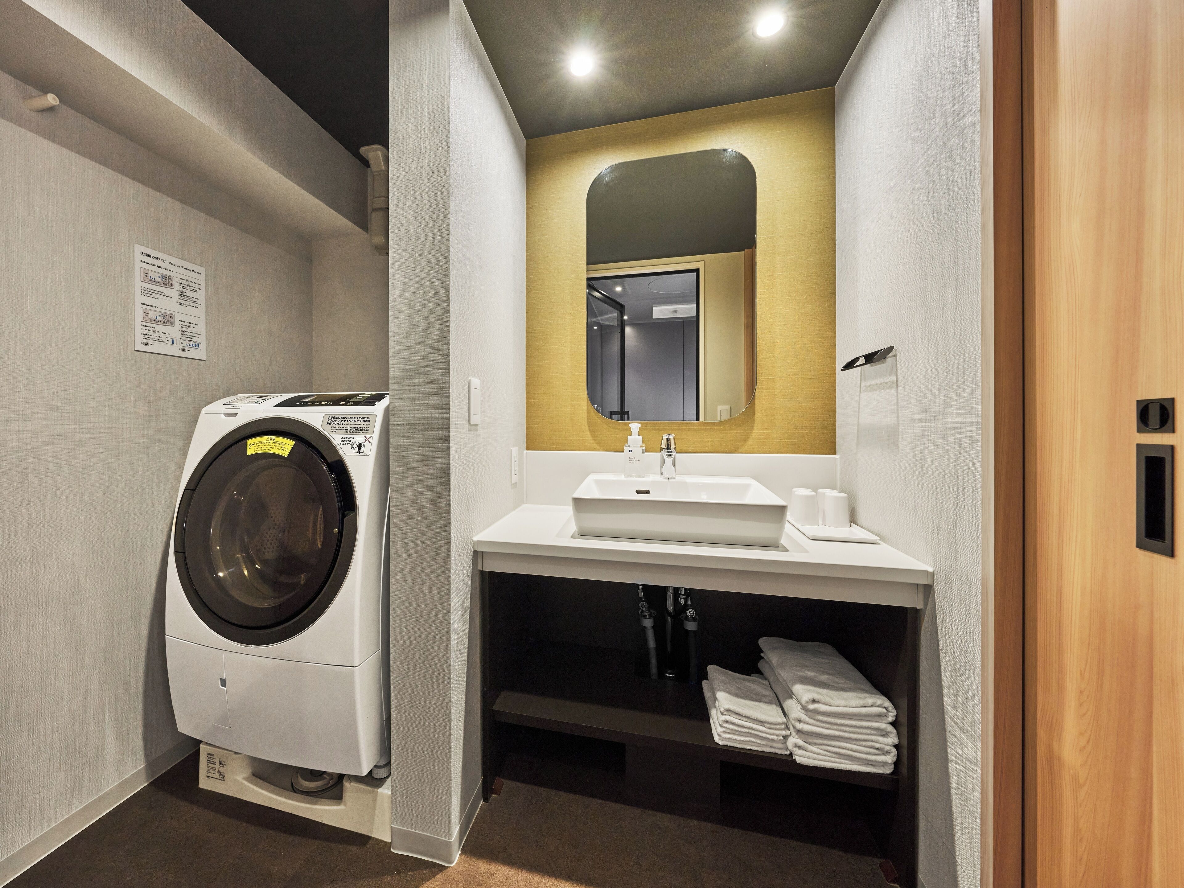 A compact hotel bathroom with a washing machine, a sink, and a mirror.