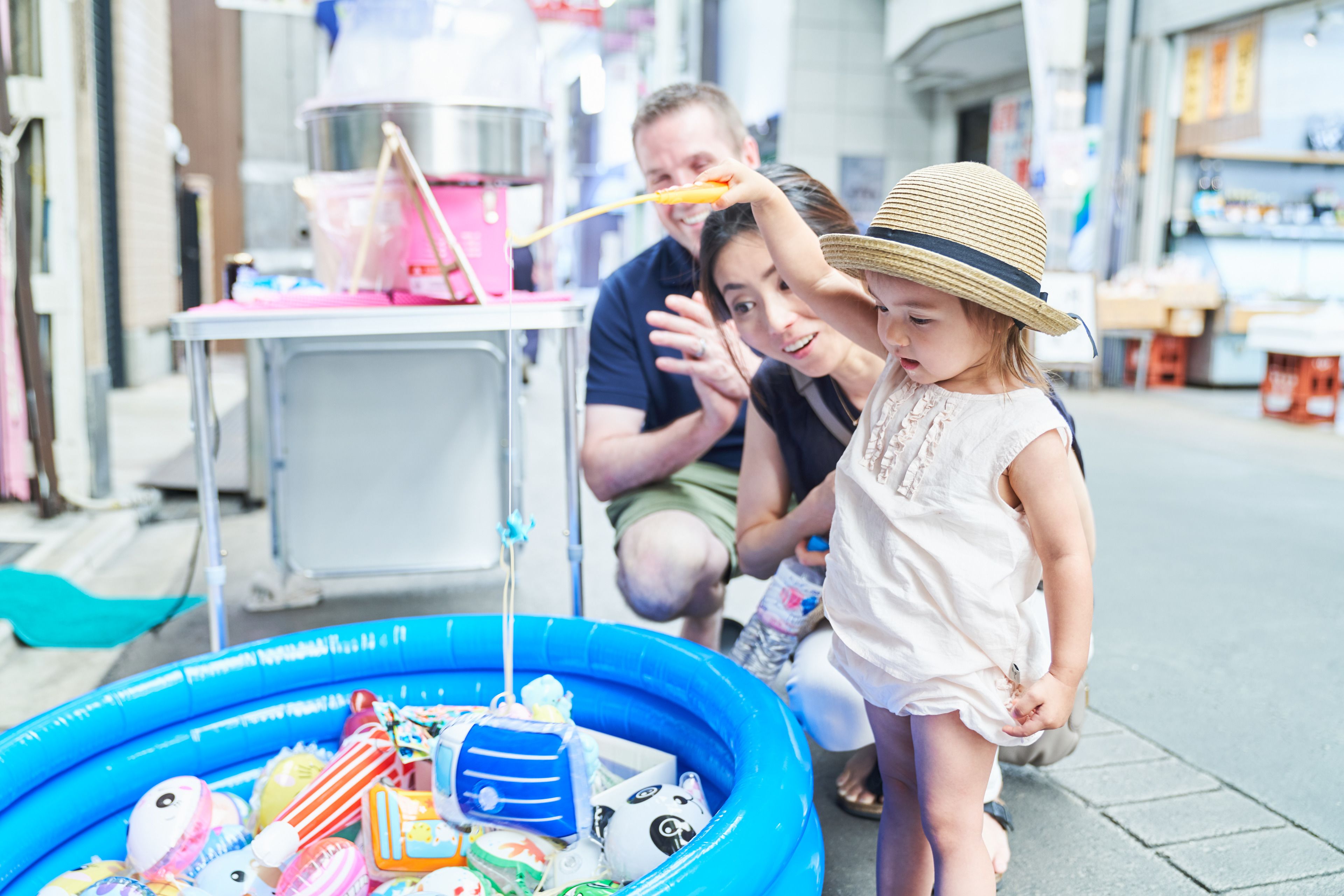 A family with a child near an inflatable pool filled with toys, a pink cooler, and a food stall in an outdoor setting.