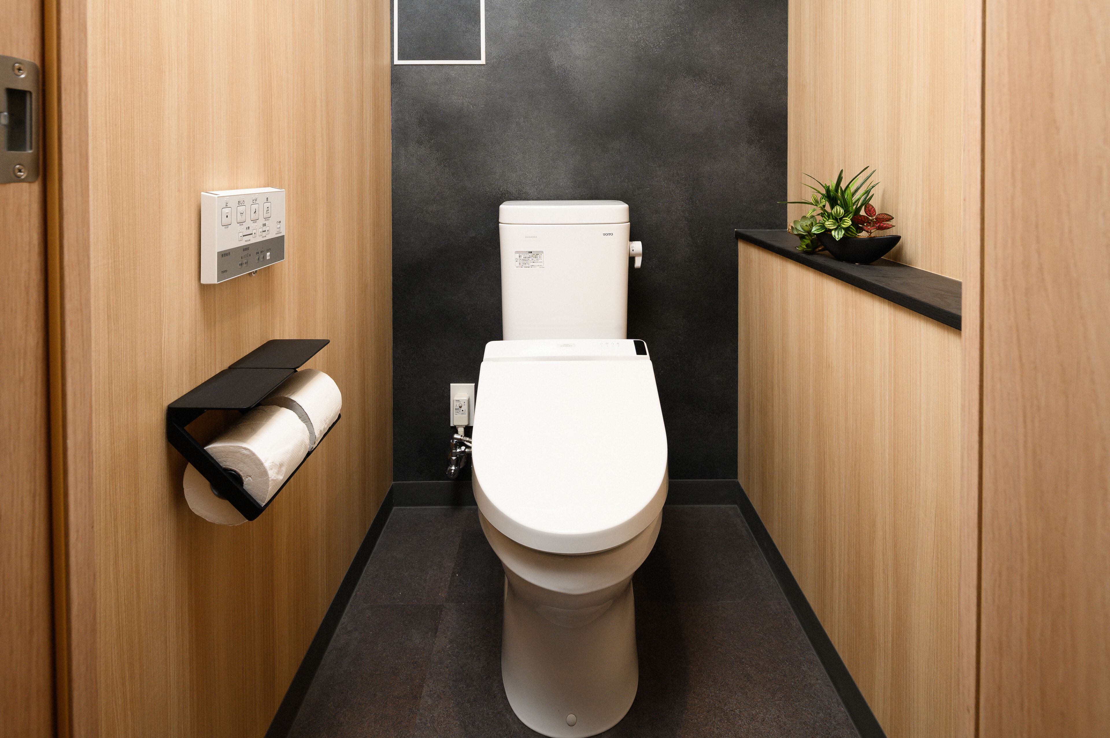 A modern bathroom with a toilet, a toilet paper holder, a black shelf, and a small plant.
