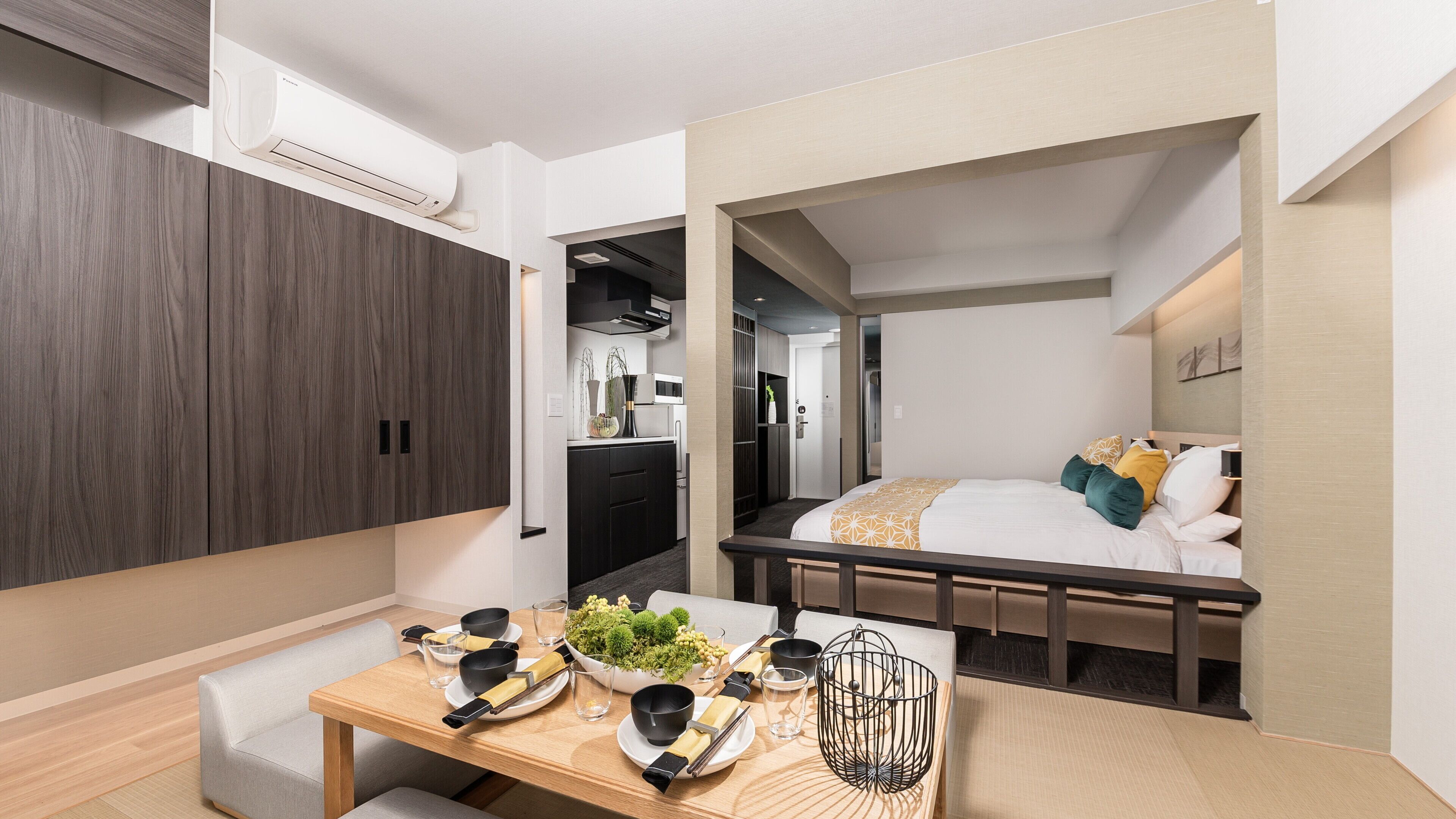 A compact hotel room with a bed, bedside table, and a kitchenette area.