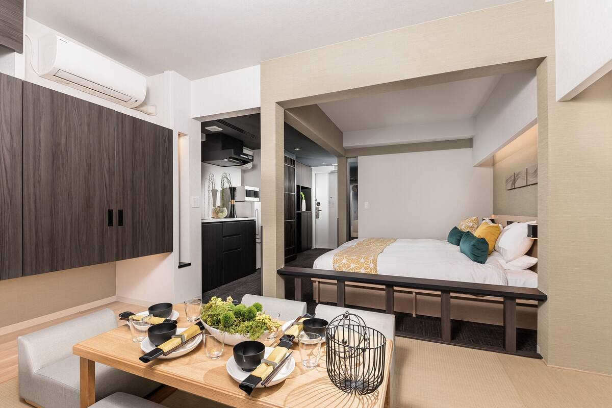 Une chambre d’hôtel compacte comprenant un coin cuisine, un lit agrémenté d’oreillers décoratifs, un coin repas avec une table dressée pour deux personnes et une grande armoire.