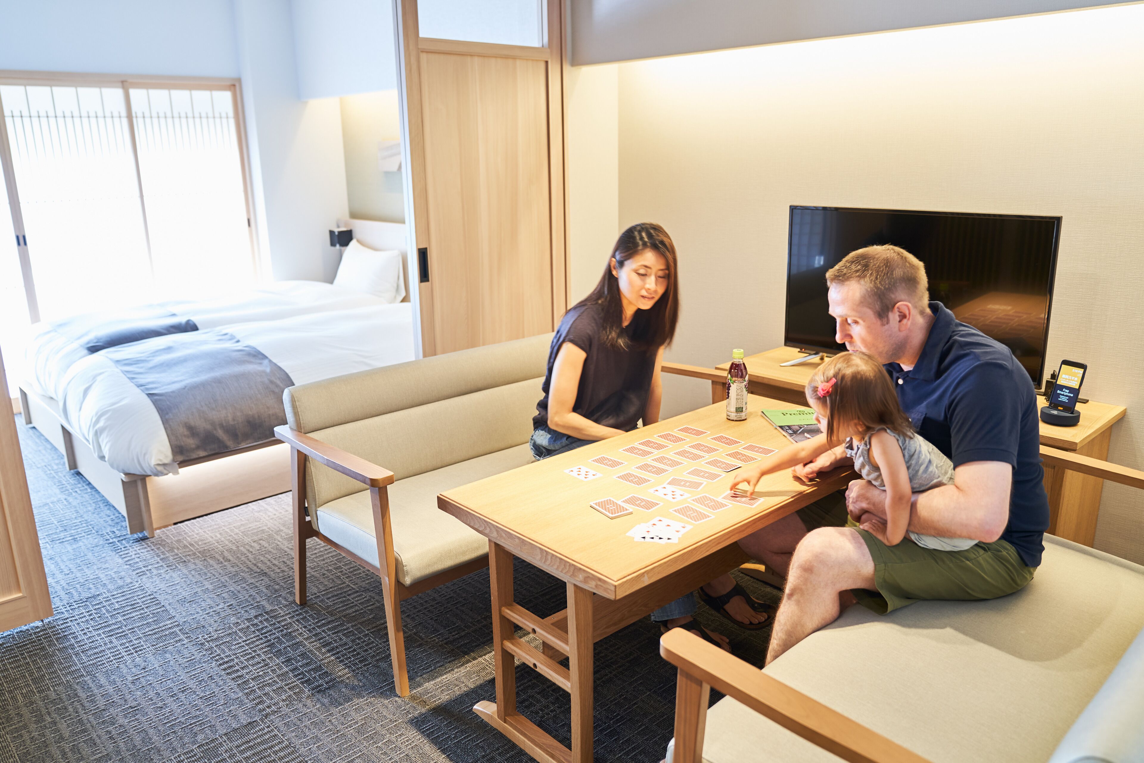A hotel room with a bed, a sofa, a wooden table with playing cards, a television, and a small table with a bottle and a remote control.