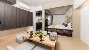 Une chambre d’hôtel moderne avec une table à manger en bois, dressée pour deux personnes, un lit recouvert d’une courtepointe blanche et dorée, et une kitchenette en arrière-plan.