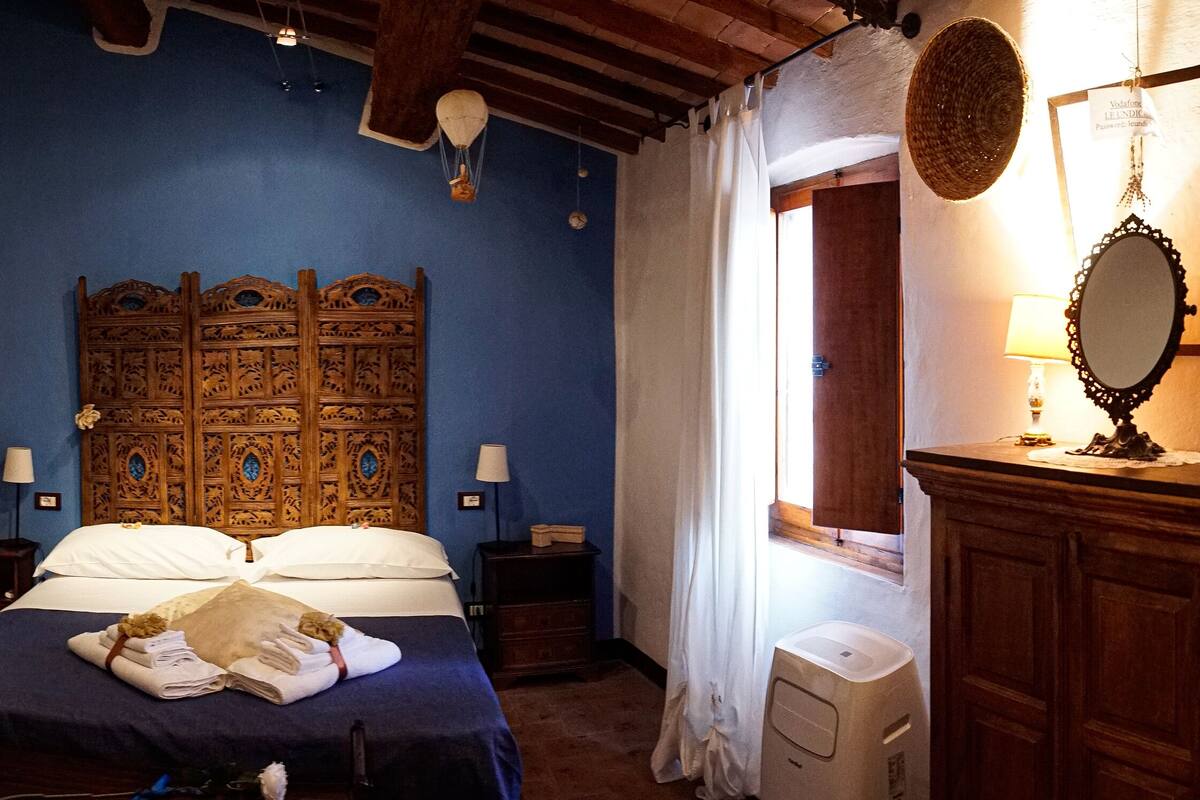 A bedroom with a wooden headboard, a bed with white linens, two bedside tables with lamps, a mirror on the dresser, and a window with curtains.