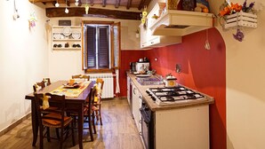 A compact kitchen with a gas stove, a dining table with chairs, and a window with blinds. - Le Undici Lune (San Gimignano)