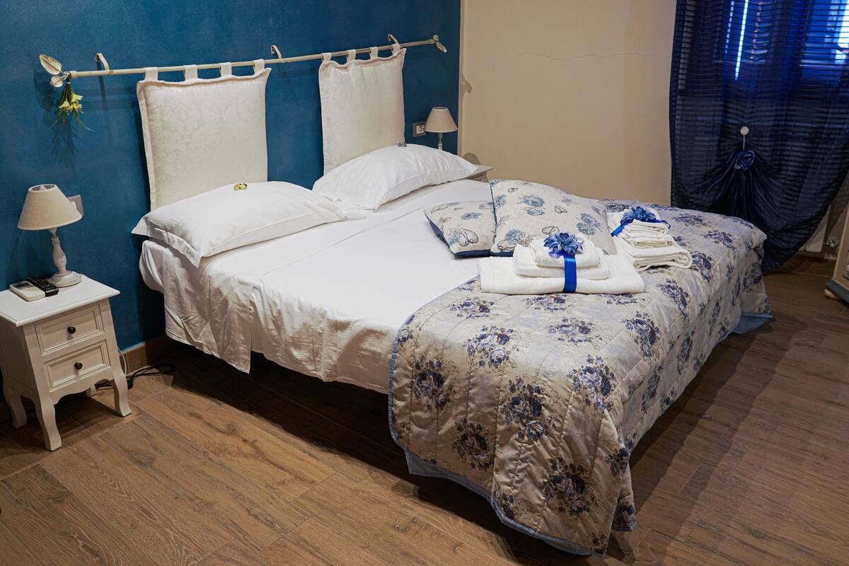 A neatly made bed with floral bedding, a bedside lamp, and a small bedside table with a lamp and a remote control.