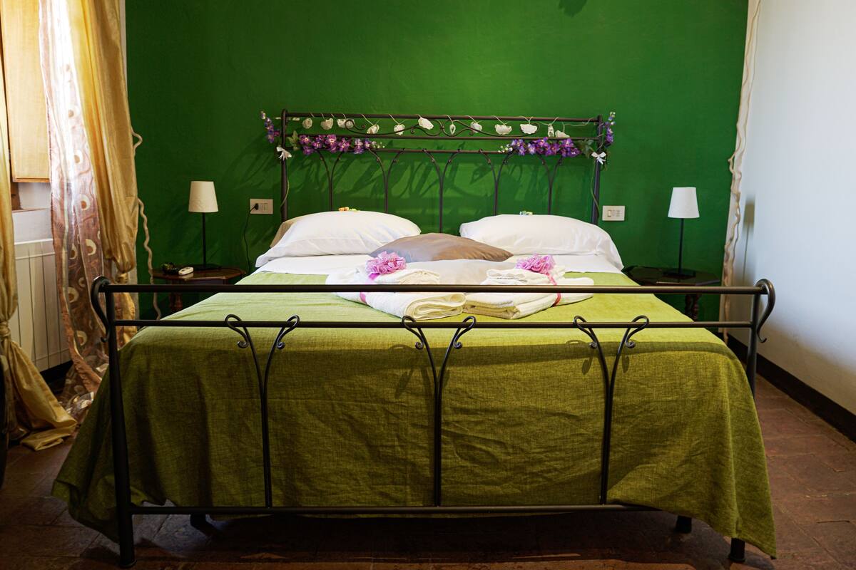 A bed with a green bedspread and white pillows, a metal headboard with floral decorations, and two bedside lamps.