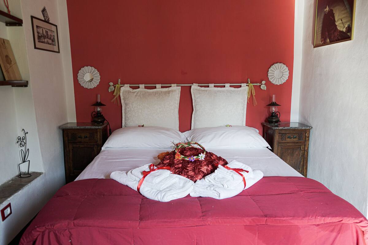 A neatly made bed with a red bedspread, white pillows, and a bouquet of flowers. There are two bedside tables with lamps and a small shelf with a plant.