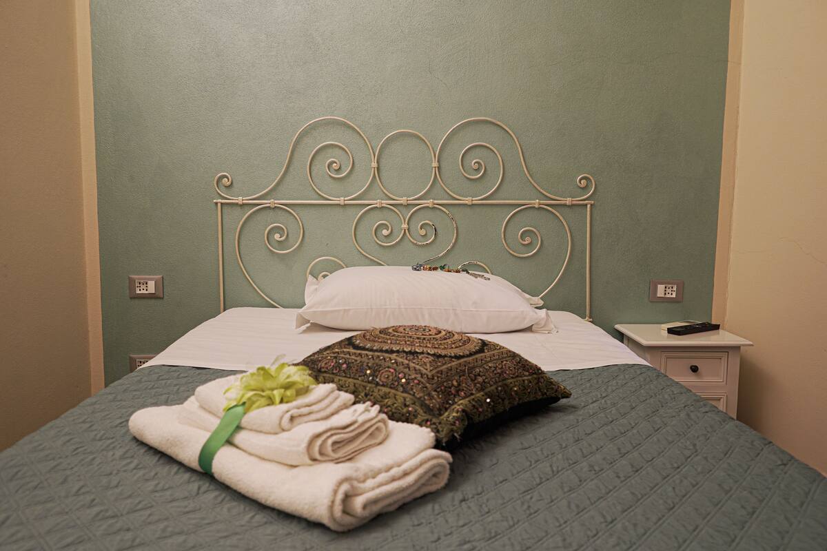 A neatly made bed with a green quilt, a white pillow, a decorative pillow, and a folded towel with a green ribbon.