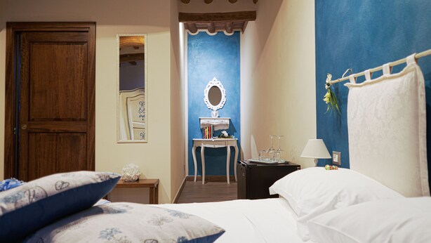 A hotel room with a bed, pillows, a nightstand with a lamp, a small table with a mirror, and a blue accent wall. - Le Undici Lune (San Gimignano)