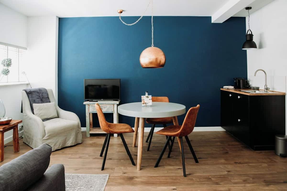 A modern living room with a blue accent wall, a round dining table with four chairs, a sofa, a TV, and a kitchen area with a sink and cabinets.