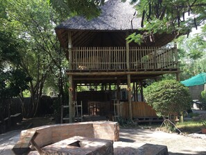 A two-story wooden structure with a thatched roof, surrounded by trees and a brick fire pit. - Laphroiag Luxury Chalets (Maun)