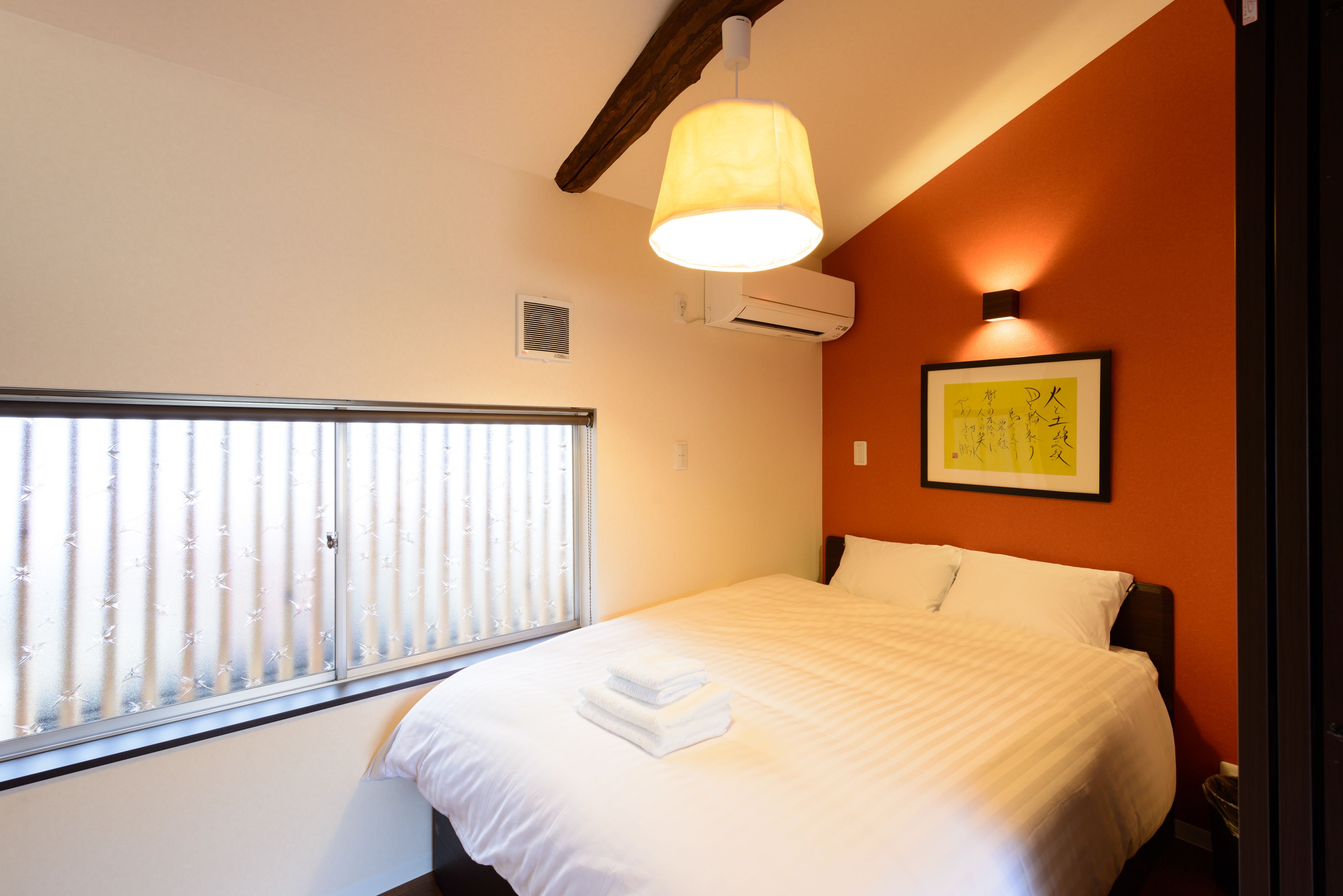 A hotel room with a bed, a window with blinds, an air conditioning unit, and a wall-mounted picture.
