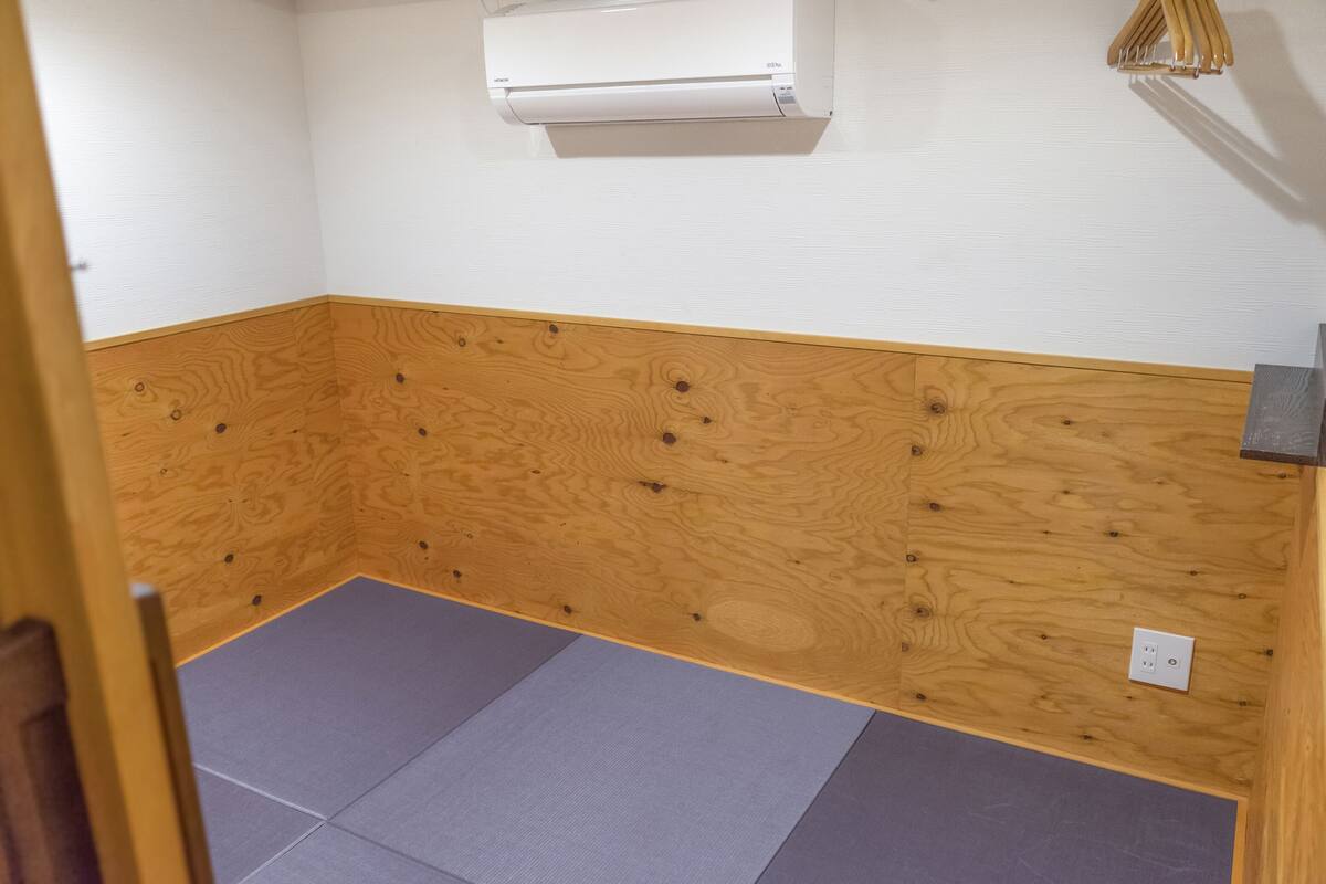 Ein Zimmer mit Holzboden, einer grauen Matte, einer weißen Klimaanlage und einem Lichtschalter an der Wand.