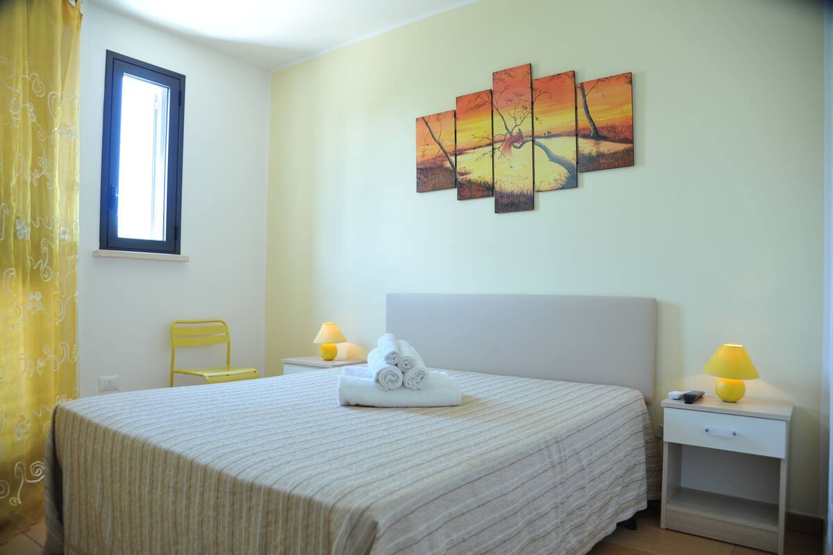 A hotel room with a bed, a window, a chair, a nightstand with a lamp, and a wall painting.