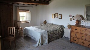 Un dormitorio con cama, una silla, cómoda, ventana con cortinas y cuadros en la pared.